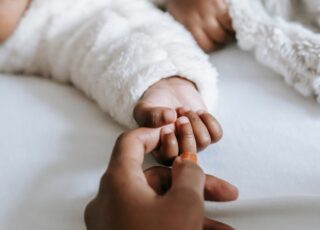 A idealização da maternidade perfeita e o peso do capacitismo. A foto mostra a mão de uma mãe, segurando a mãzinha de seu bebê. Ambos são pretos.