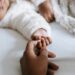 A idealização da maternidade perfeita e o peso do capacitismo. A foto mostra a mão de uma mãe, segurando a mãzinha de seu bebê. Ambos são pretos.