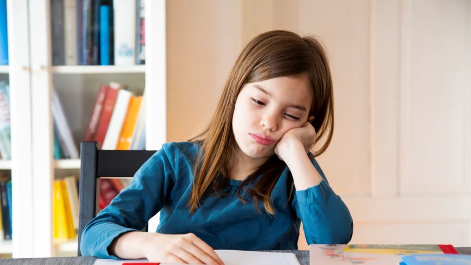 Garota adolescente, sentada à mesa, com livros e cadernos, à sua frente. Em seu rosto jovem, a expressão de aborrecimento.